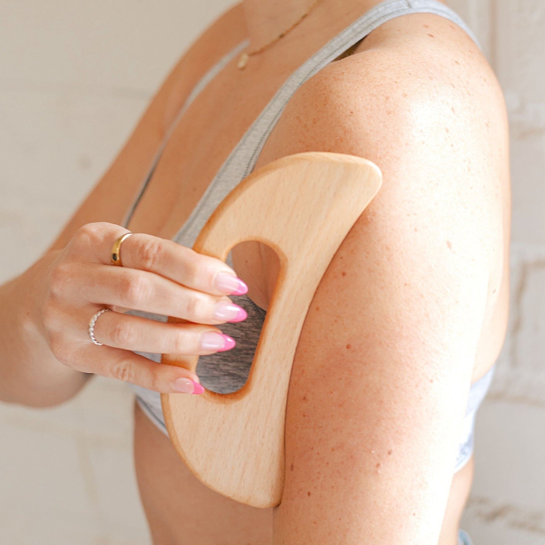 A woman using a wooden lymphatic drainage tool on her left arm while her right hand holds the tool against her skin. She is wearing a gray sports bra standing in front of a white brick background.