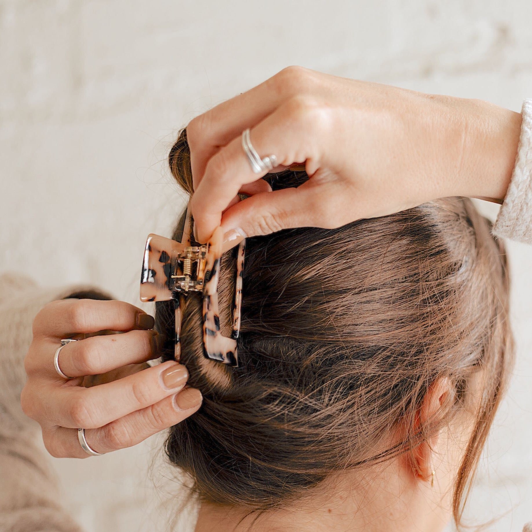 A girl with brunette hair wearing a beige tortoise rectangle claw clip in her hair in a updo. She is standing in front of a white bright wall.