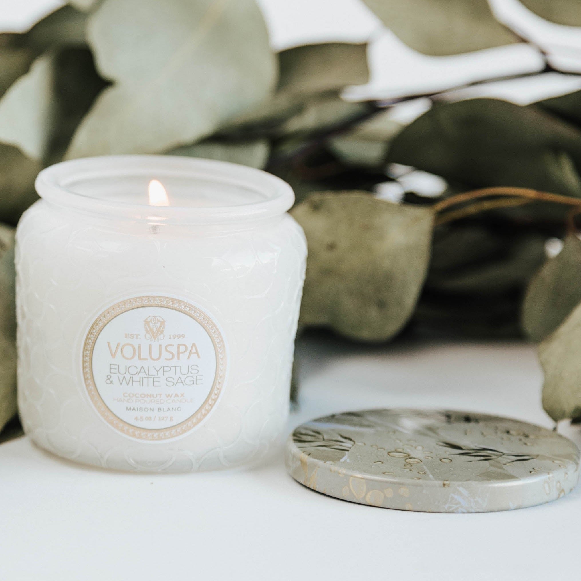 Lit candle in white glass vessel surrounded by eucalyptus.