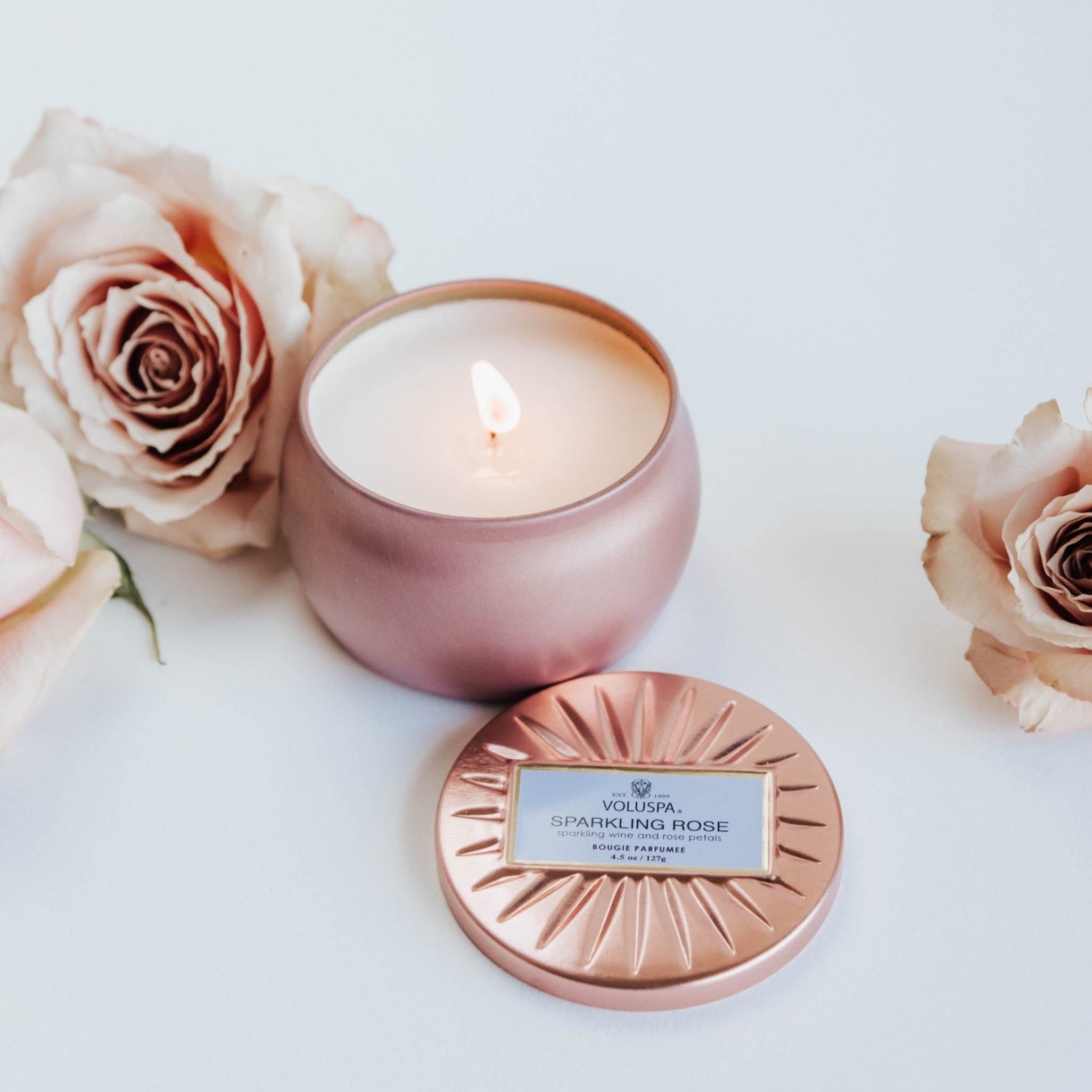 Pink sparking rose tin candle lit next to dusty pink roses.