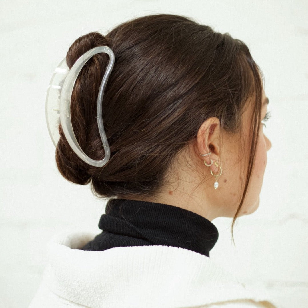 A woman with brunette hair pulled into an updo, secured with a clear acrylic half moon claw clip. She is standing in front of a white brick wall.