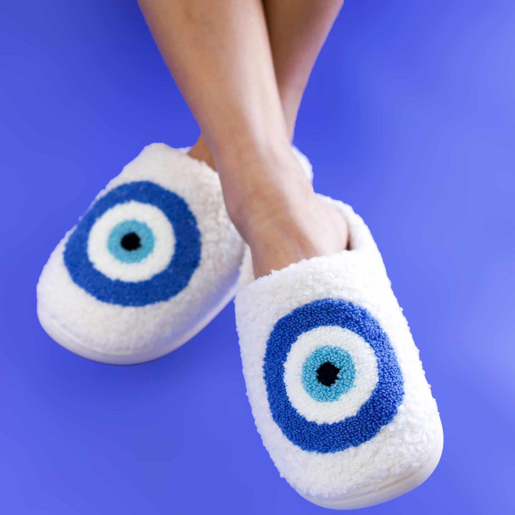 A woman's legs/feet wearing a pair of blue, white, and black evil eye slippers on a cobalt blue background.