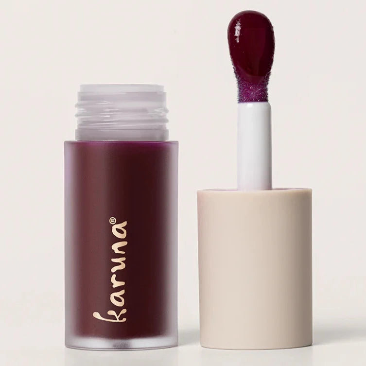 A purple colored lip oil that reads, "KARUNA." The doe-foot applicator is displayed next to the lip oil.