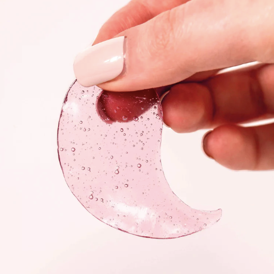 A caucasian hand with beige painted nails holding a single Rosé Hydrating Under Eye Gel. Photographed on white background.