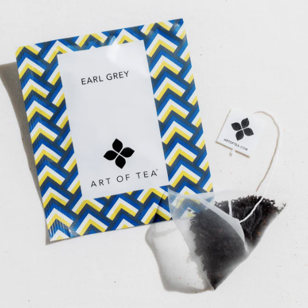 a clear tea sachet with white Art of Tea Label next to tea bag packaging with blue and yellow geometric design