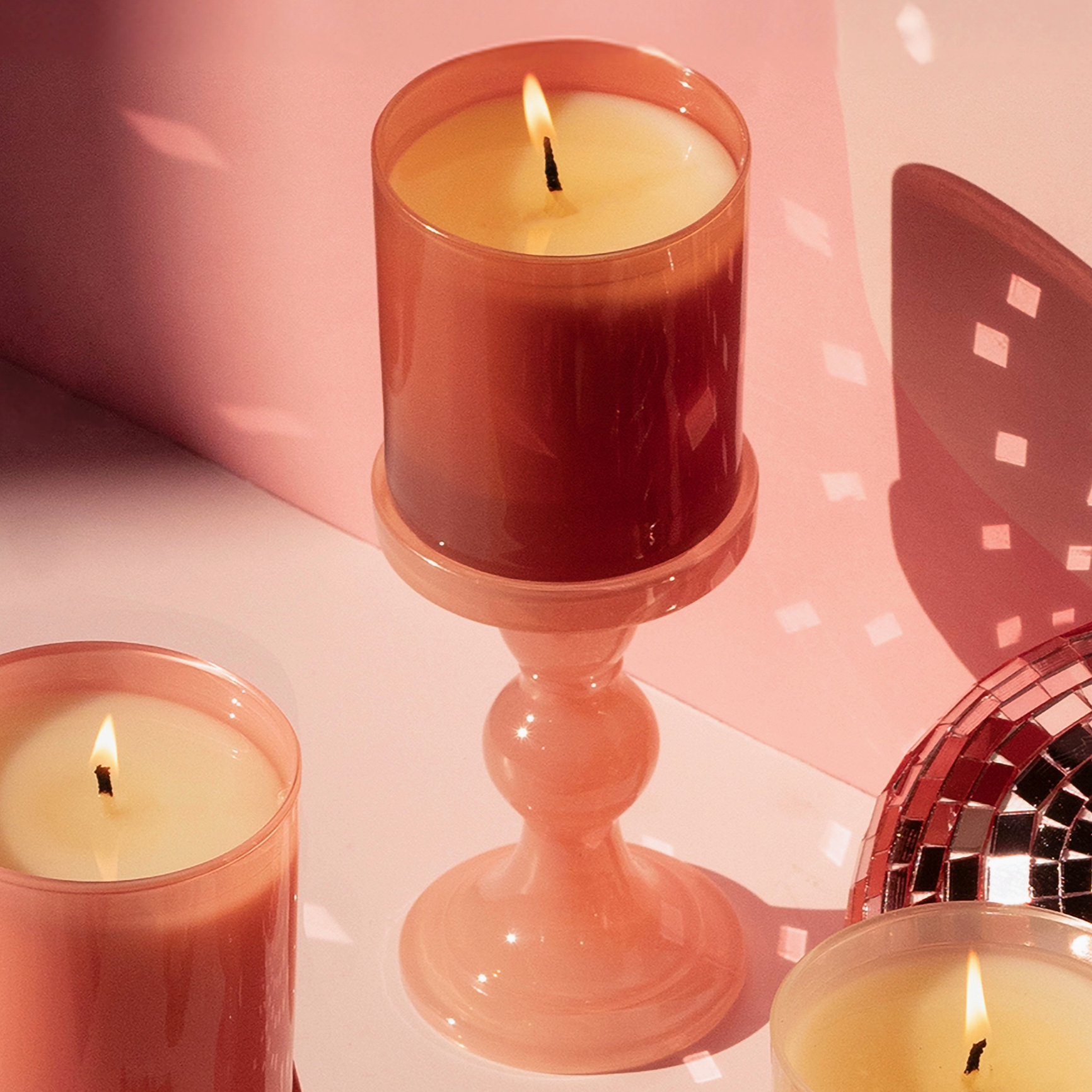 candle burning in amber glass on holder behind pink background next to disco ball and other candles