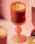 candle burning in amber glass on holder behind pink background next to disco ball and other candles