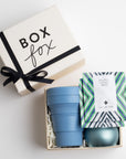 Creme BOXFOX gift box with a bow, next to an open gift box containing a blue collapsible cup, a blue tin candle and two art of tea sachets.