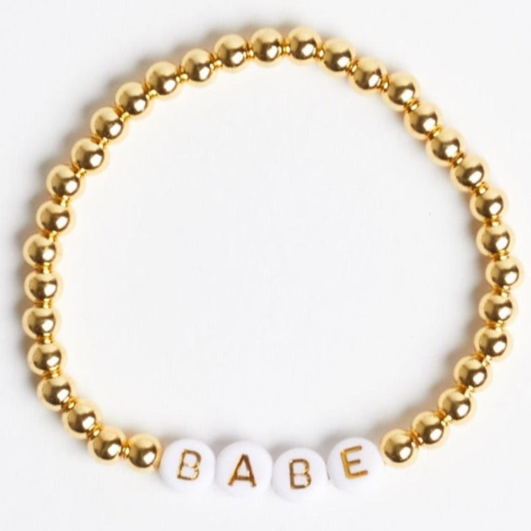 A gold beaded stretch bracelet with white beads that have gold text reading "B" "A" "B" "E" to spell out "BABE." Photographed against a white background.